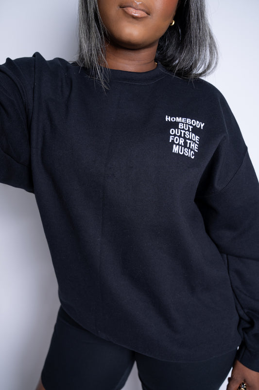 Woman wearing black embroidered sweatshirt with 'Homebody but outside for the music' stitched on the left chest