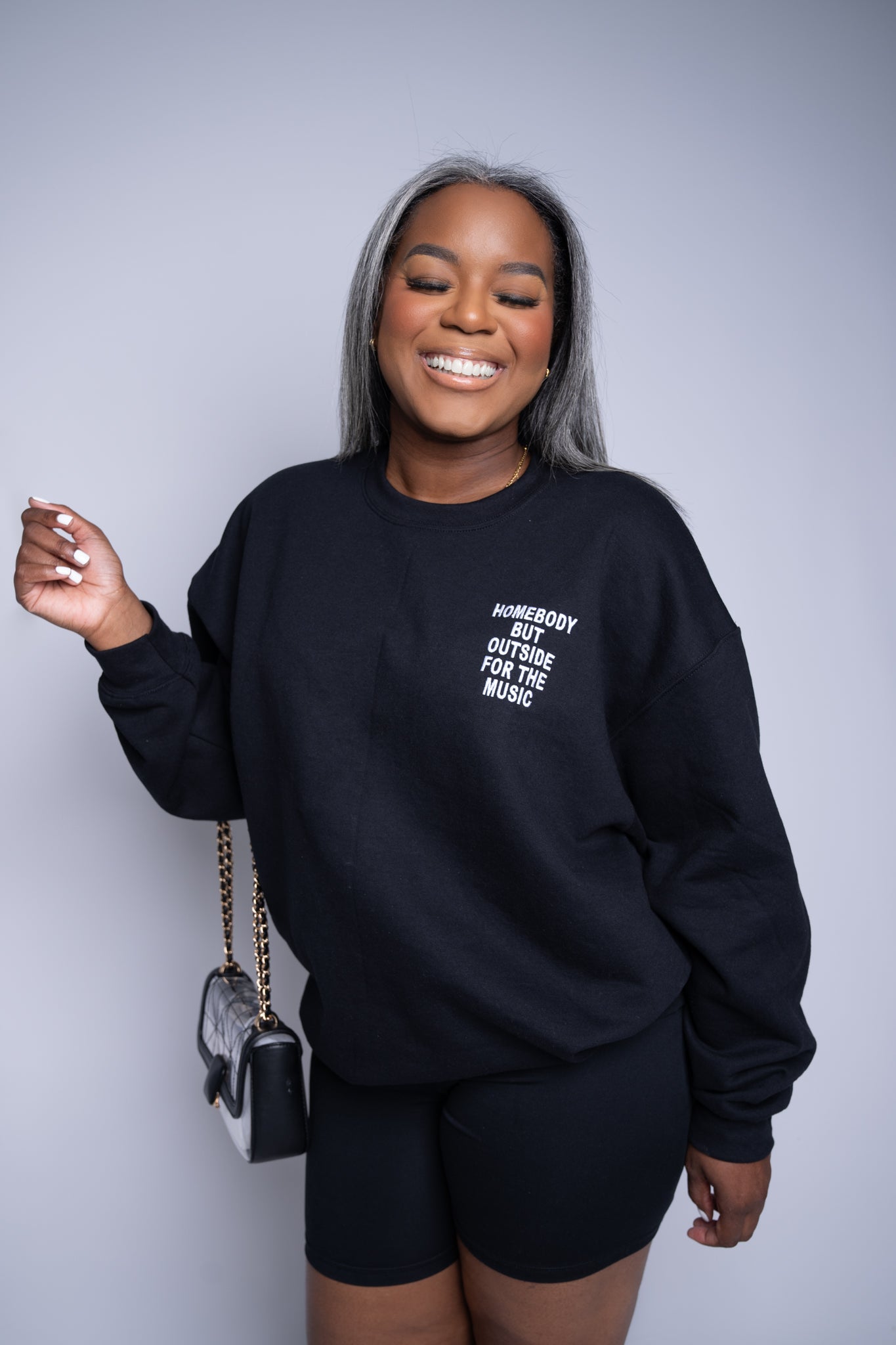 Woman carrying a purse, wearing black embroidered sweatshirt with 'Homebody but outside for the music' stitched on the left chest